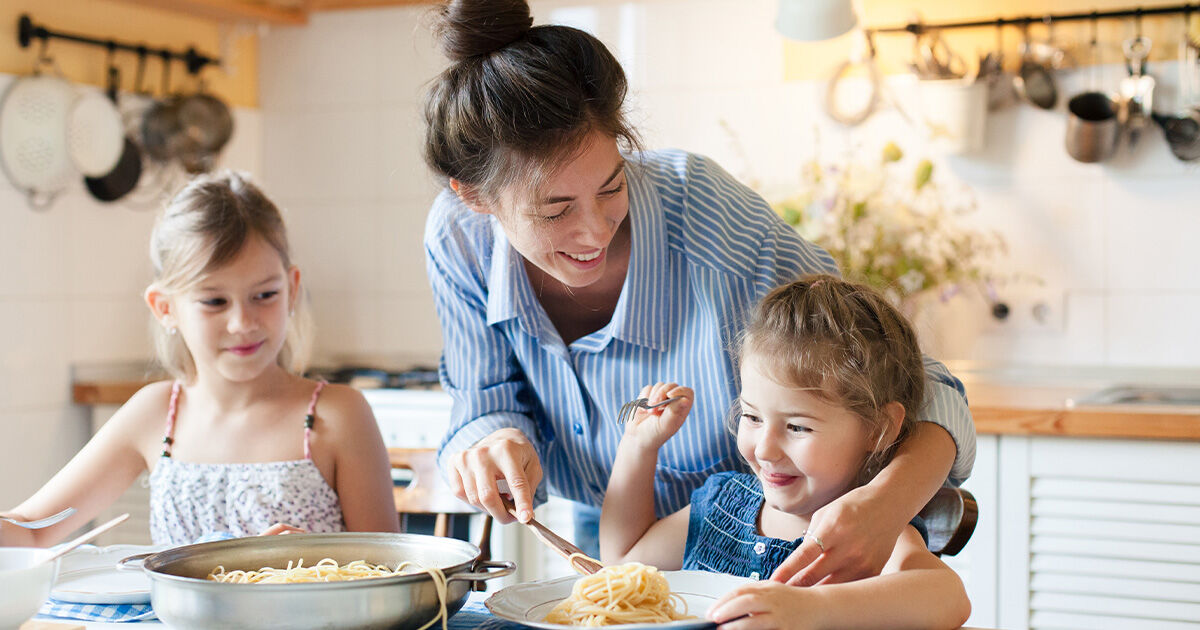 CC-Community-Header-3 Parent serving pasta to two young children seated at a kitchen table during a family meal.
